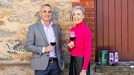 Jason Spiteri and Carmen Houston posing in front of a building, holding glasses of red wine.  Two boxes of wine are on the steps.