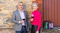 Jason Spiteri and Carmen Houston posing in front of a building, holding glasses of red wine.  Two boxes of wine are on the steps.