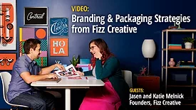 Fizz Creative founders, Jason and Katie Melnick are in a room seated opposite each other on a white table looking at designs, a white lamp and shelf to the right. The purple wall is decorated with varying sizes of typography and design.