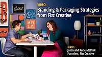 Fizz Creative founders, Jason and Katie Melnick are in a room seated opposite each other on a white table looking at designs, a white lamp and shelf to the right. The purple wall is decorated with varying sizes of typography and design.